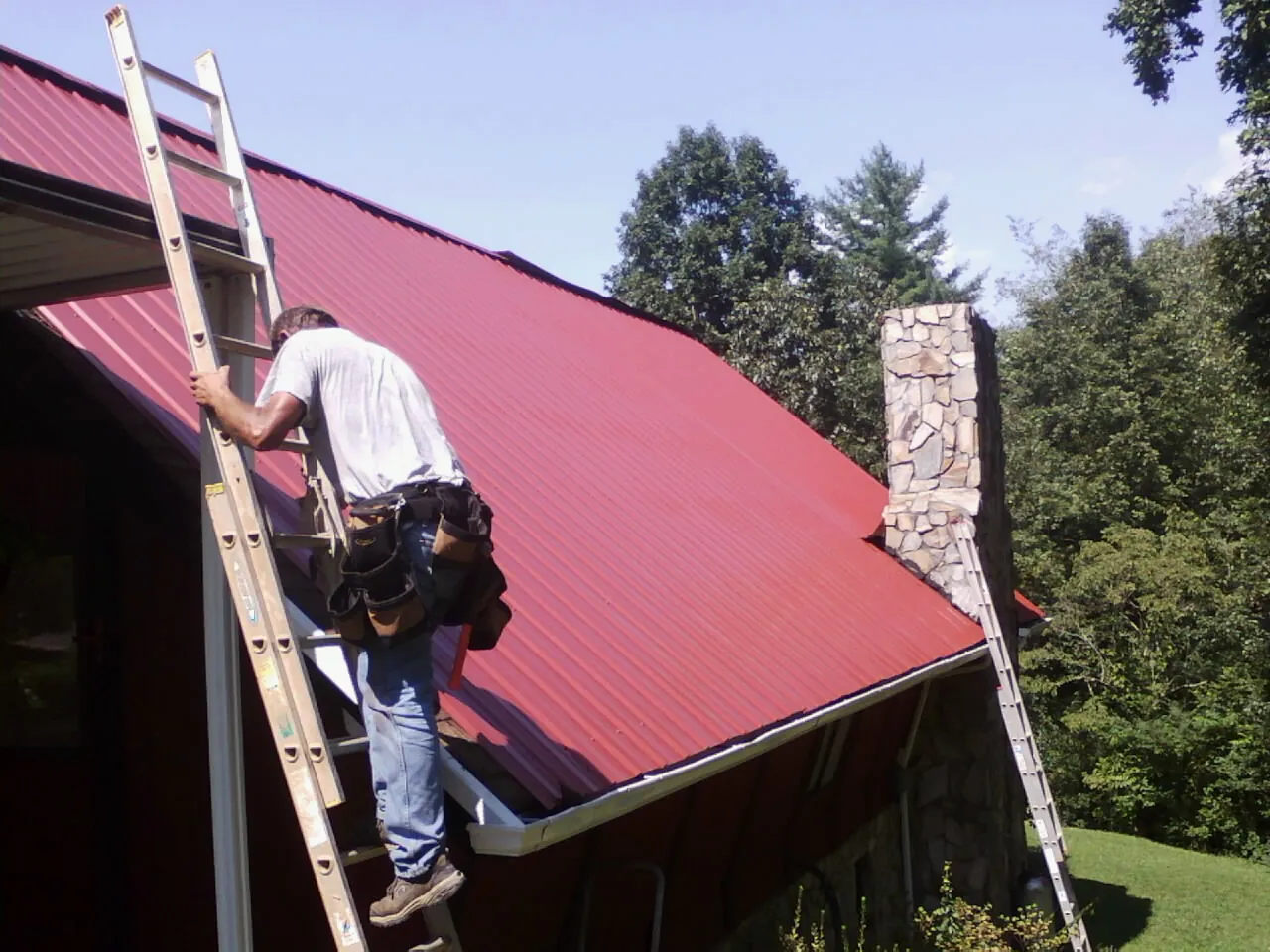 Roofer on a ladder beside a red metal roof and stone chimney — Soffit & Fascia Repair by Roofing Brothers in Micro